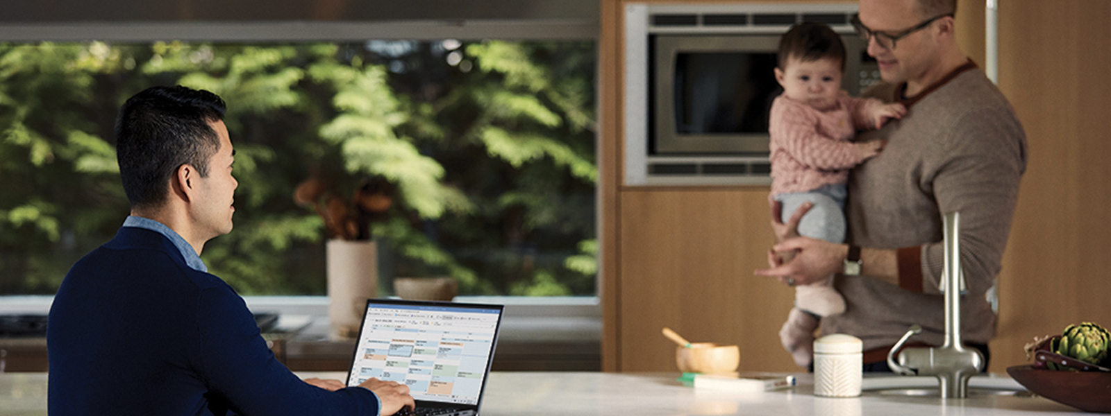 a father holding his baby in the kitchen while talking to a man using a laptop.