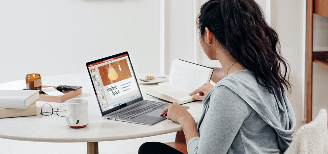 A student sitting at a table referencing a book while building a PowerPoint presentation.
