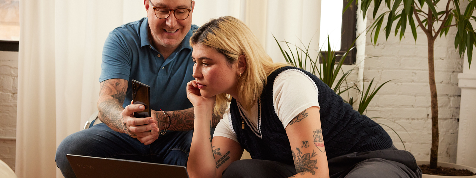 Two tattooed people looking at a laptop and phone sharing scholarship opportunities.