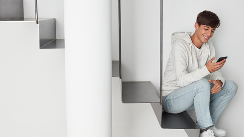 a male student inside home, smiling, and looking at his phone.